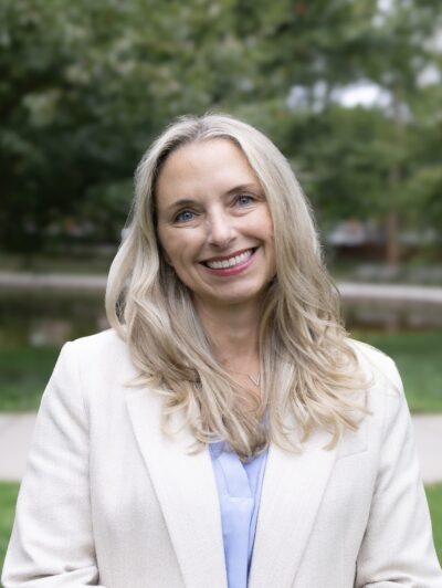 A smiling woman in a cream suit standing outside with foliage behind