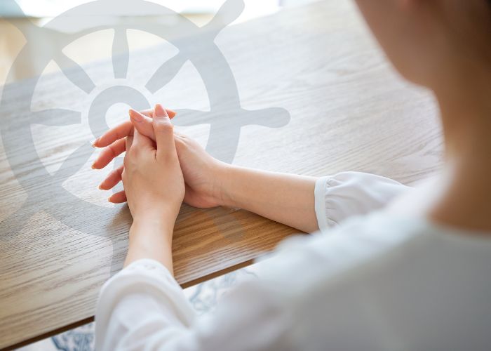 A woman sitting at a table with her hands folded; a semi-transparent ship's steering wheel is layered over the image.